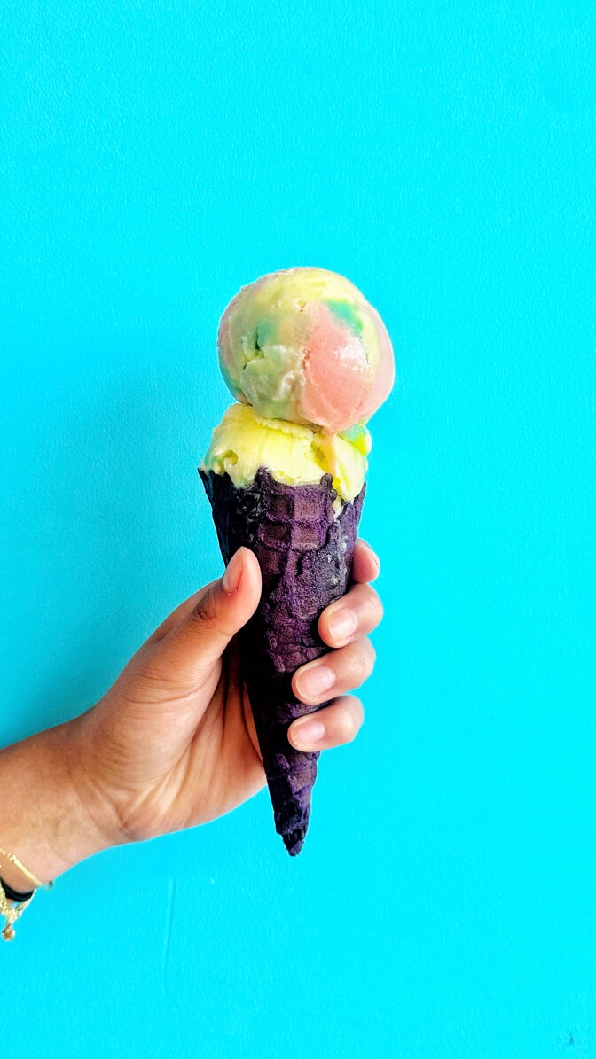 A double scoop of pastel green, yellow and pink swirled sherbet in a purple waffle cone, held up against a bright teal background.