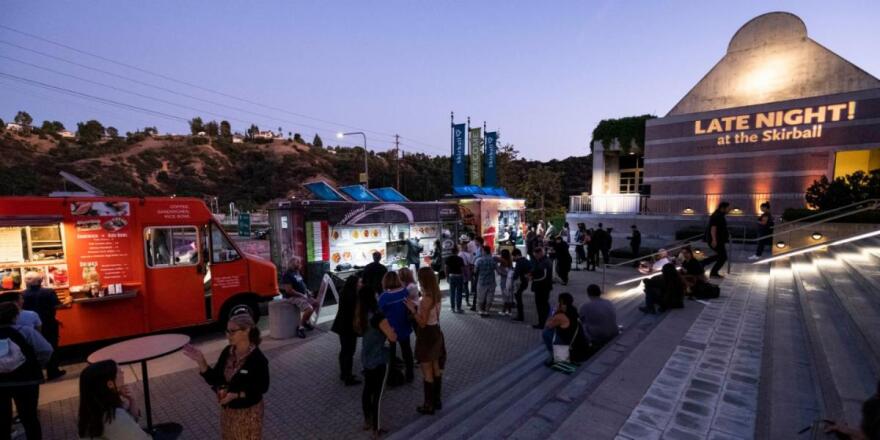 The front steps and building of The Skirball Center with food trucks in the lane and people standing and sitting on stairs.