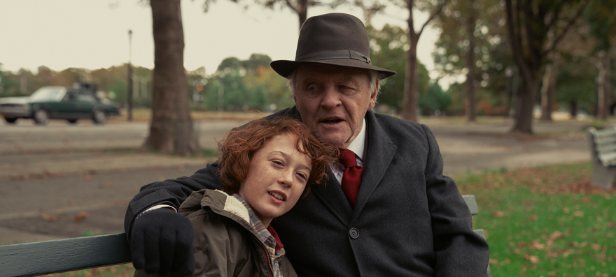 Anthony Hopkins and Banks Repeta sit together on a bench in a park, an otherwise empty street behind them except for one green car in the left. Hopkins wears brown fedora and a dark grey coat. The coat is buttoned, allowing for only the collar of a white button up with a red tie to be visible underneath. Hopkins has his right arm around Repeta, his black gloved hand resting on Repeta's shoulder. Hopkins' chin rests on Repeta's head, as Repeta leans against Hopkins chest, his reddish curly hair a uncombed. Repeta wears an army green jacket, a grey flannel peaking out slightly from underneath. Repeta looks out at the park as Hopkins looks off to the left, his mouth open as if in mid conversation.