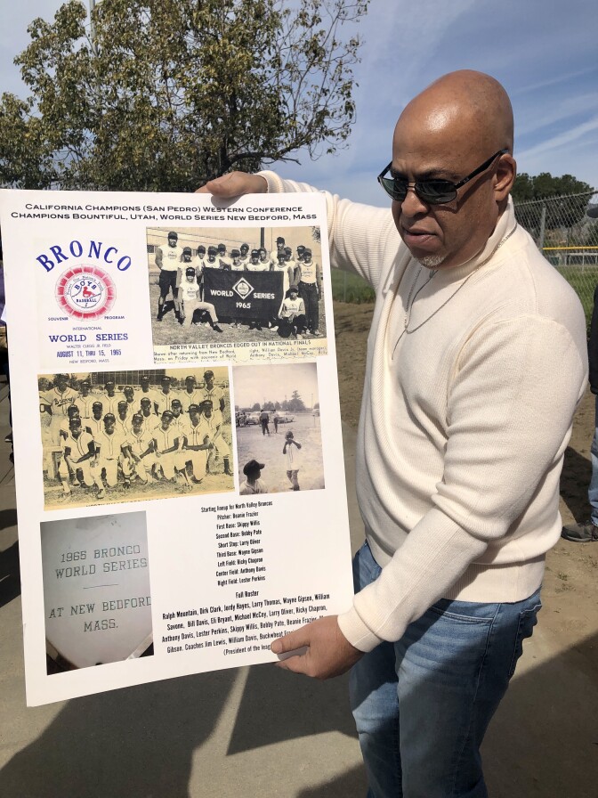 Black man with sunglasses and white long sleeve shirt holds posterboard with black and white photos