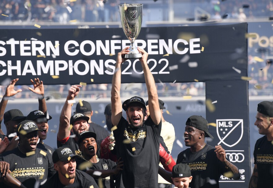 A group of men with a range of skin tones cheer and celebrate as one man hoists an MLS trophy over his head. Sign behind them reads: Wester Conference Champions 2022.