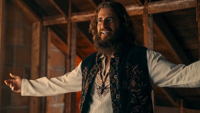 Johnathan Roumie stands in a barn-like building with wooden rafters visible behind him. Roumie's arms are raised almost shoulder height and are outstretched to his sides. He wears a loose white long sleeve that has a v-neck embroidered with black and orange filigree. Perfectly lined with the v-neck collar of his short hangs a beaded necklace with a cross pendent. Over the shirt is a black vest that has gold embroidered flowers across the chest. Roumie smiles largely. He has a long scraggly beard and shoulder length hair that falls in loose waves. He is portraying the American Evangelist Lonnie Frisbee who was a key figure in the Jesus Movement of the 1960s and 1970s.