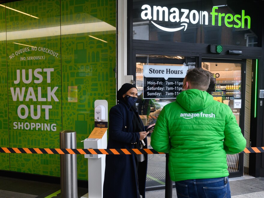 A person in a green Amazon Fresh puffer jacket speaks to a customer near a "Just Walk Out shopping" sign.