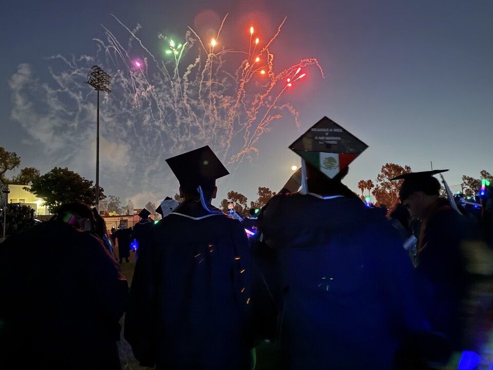 Students in graduation regalia are seen from the back, looking up at fireworks in the night sky.
