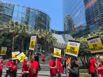 Protesters of varying skin tones wearing red shirts are carrying signs that read "ON STRIKE" outside in downtown Los Angeles. 