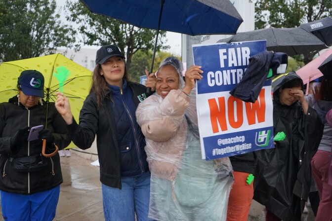 Two people stand under an umbrella smiling. One of them holds a sign that reads "Fair Contract NOW." One person with long brown hair and medium-light skin tone wears a blue hat. The other person with medium-dark skin tone wears a pink jacket and poncho.