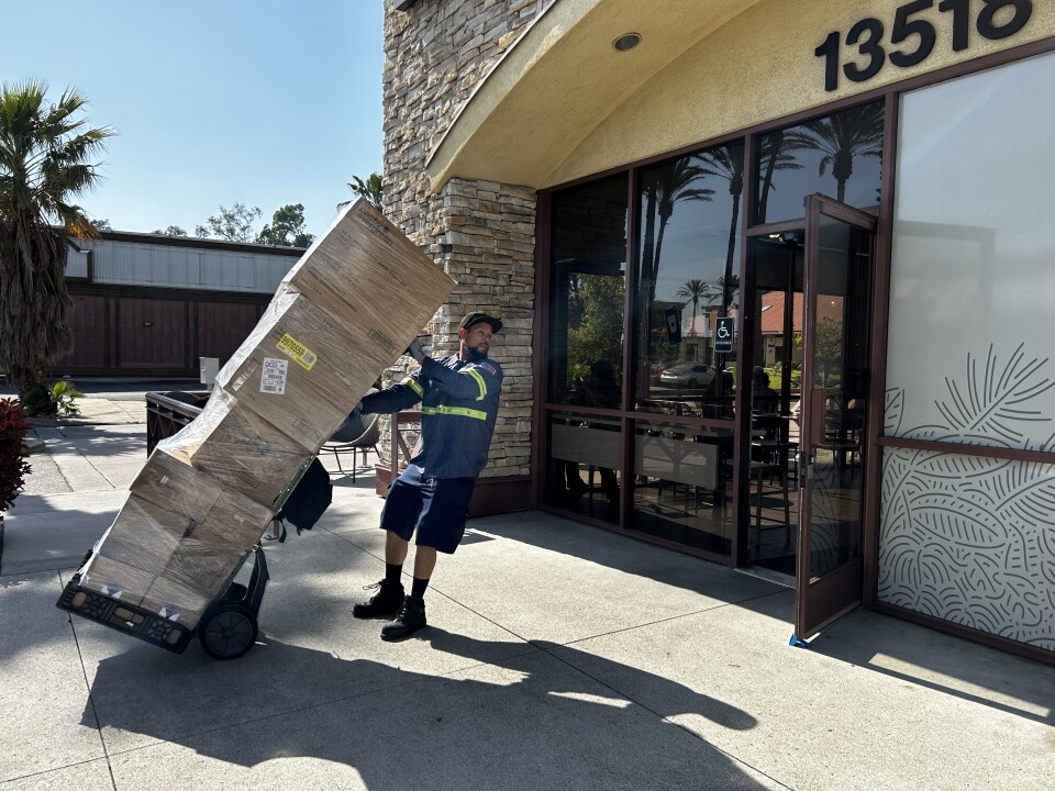A man pulls a large stack of boxes on a dolly, backing into the entrance of a storefront. 