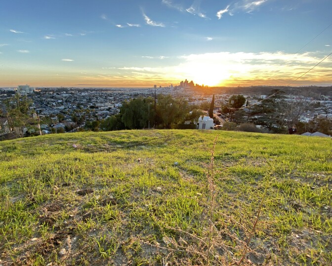 A wide view from the grassy hilltop at sunset. The cityscape of downtown Los Angeles is viewable in the distance.