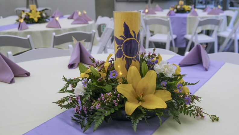 A floral centerpiece sits on a table with a purple table runner and purple napkins. Yellow lilies, purple asters, white carnations and pink waxflower surround a yellow lantern with a purple sun design.