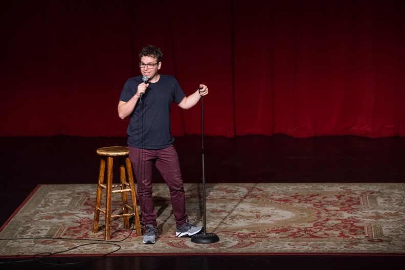 Comedian Jon Lovett is shown onstage, holding a microphone and doing a comedy set.