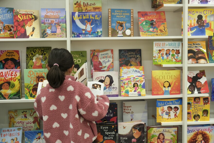 An 8-year-old child wearing a pink sweater with white hearts with with medium light skin tone browses picture books featuring characters with a variety of skin tones at a children's bookstore.