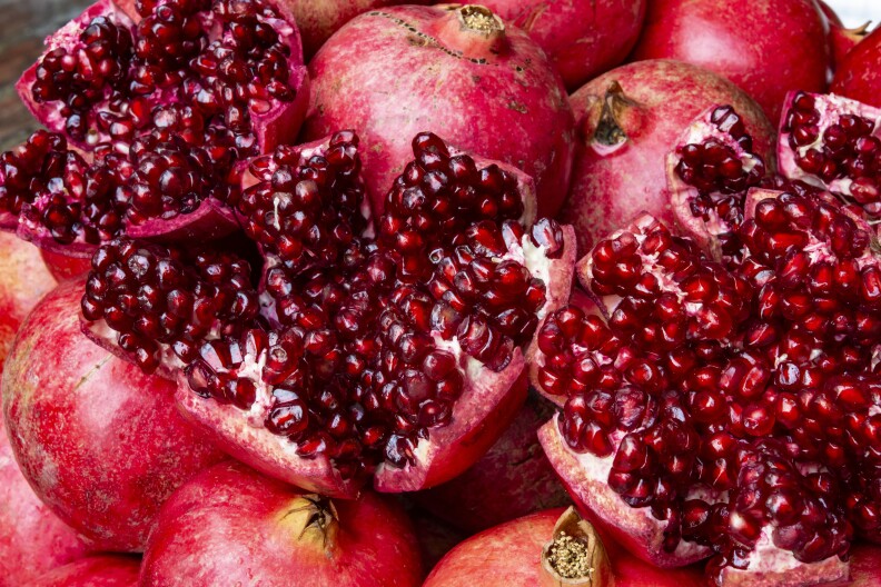 A close-up look at more than a dozen ripe pomegranates all piled upon each other: A few of the fruits have been broken open to expose the interior, showcasing all the shiny, glossy seeds inside, ready to eat.