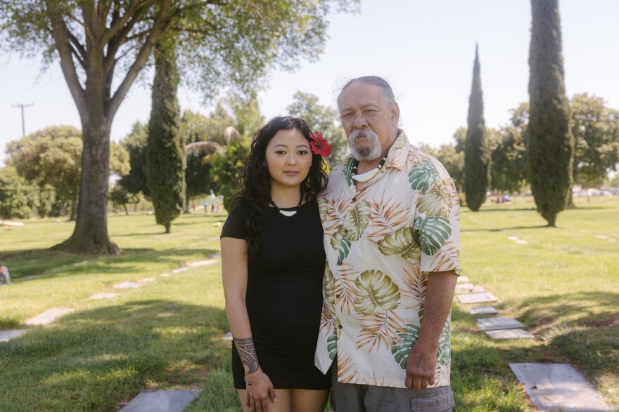 An older man with medium-light skin tone and a gray goatee wearing a shirt with tropical plants stands next to a young woman with medium-light skin tone long dark hair and a hibiscus flower in her hair. They are standing in the lawn of a cemetery.