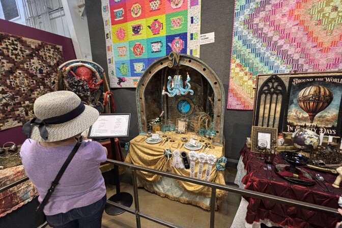 A woman in a purple t-shirt and gray straw bucket hat with black bow looks at table setting displays. In the center is an ocean-themed table with a blue octopus chandelier, gold draped tablecloth, and a porthole in the background.