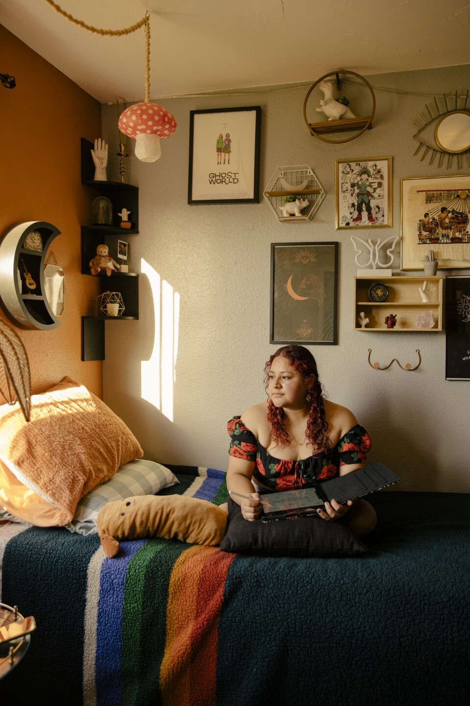 A woman with reddish-brown, curly hair sits cross-legged on a bed with sun coming through a side window. She's wearing a black off-the-shoulder blouse printed with large red flowers and she's holding a laptop that opens flat. 