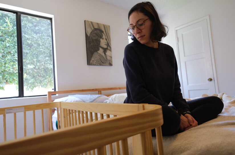 A woman with medium-tone skin and glasses looks down on a wood crib while sitting on a bed. A photo of a woman hangs on a white wall between windows.