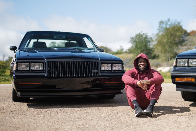 Kevin Hart, in red sweats and a red hoodie sweatshirt, sits on the ground next to his custom black Buick.