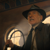 Harrison Ford stands in a large room that seems to be inside a temple or church of some sort. The walls are made of sand colored stone with large windows of curved shapes. The windows let warm light in from the outside. Ford wears the classic Indiana Jones outfit: a brown hat, brown leather jacket, whit button-up and black tie. He raises his iconic whip in his right hand, bracing to use it. His face looks like one of shock or surprise, as if caught off guard by whoever stands in front of him out of view. His eyes are wide, his mouth slightly open but tensed. 