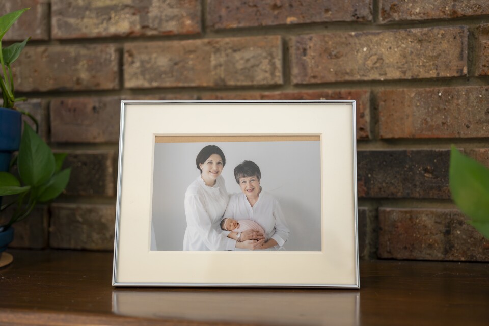 A framed photograph of two women, both with light skin tone wearing light-colored clothes in front of a white background, pose for a photo where one woman is holding a baby wrapped in a pink blanket. The frame sits on a wooden shelf in front of a brick wall.
