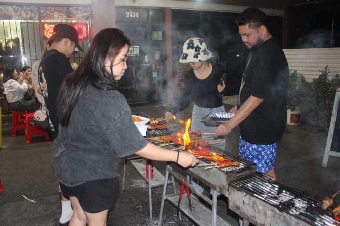 People grilling skewers over open flame on charcoal grills set up on a sidewalk at night, with smoke rising and red plastic stools visible in the background.