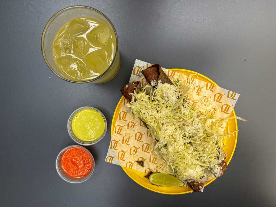 A plastic yellow plate holds a to-go wrapper with a golden-lettered design. It is topped with two large flautas made from rolled blue corn tortillas, shredded cabbage, and a small wedge of lime. To the left of the plate, two small plastic containers are filled with red and green salsa. Above the plate is a glass containing a pale greenish-yellow liquid.