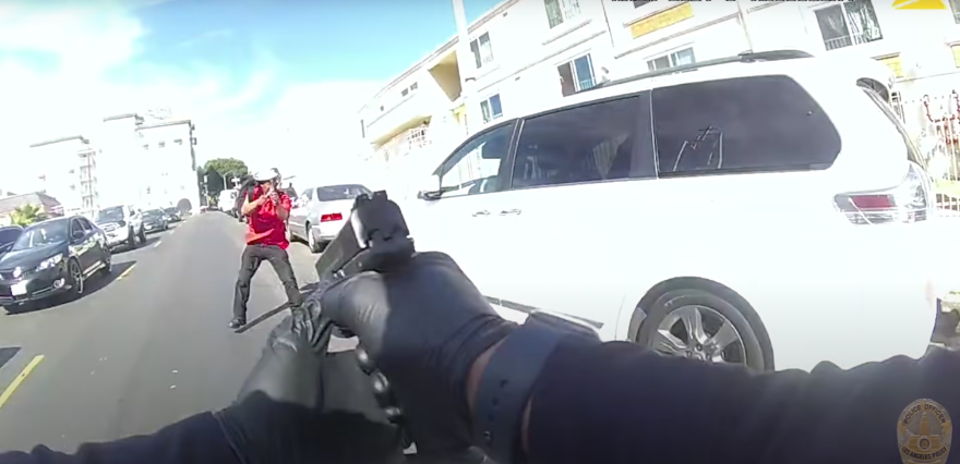 In a police officer's point of view, a pair of hands wearing black gloves grip a pistol aimed ahead at a man wearing black pants and a red shirt who seems to be aiming something back at the officer. The man is standing in the road with a line of cars in traffic on one side and white van parked on the other.