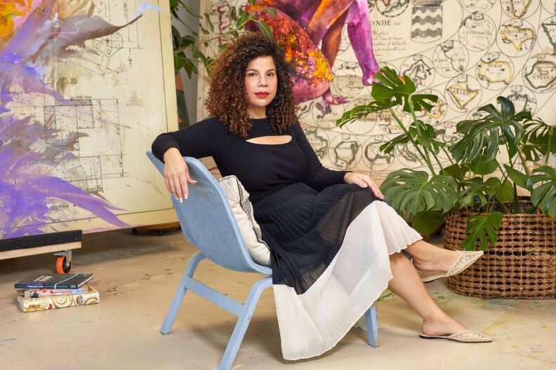A portrait of artist Firelei Báez. She's sitting on a blue chair, wearing a black and white dress. Behind her are two large canvasses of artwork. A green plant is in a weaved basket to the right of the photo.