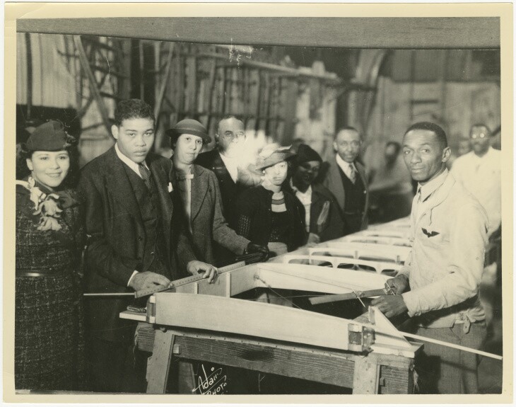 several Black men and women stand around the frame for a partially built airplane wing.