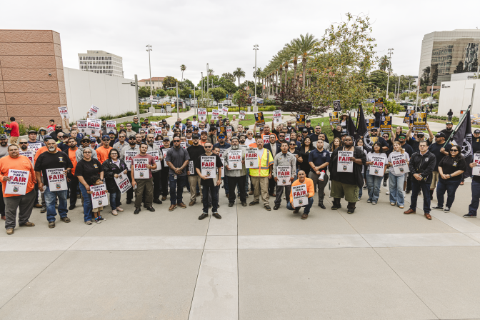 A group of people holding union signs and flags stands in front of a government building. 
