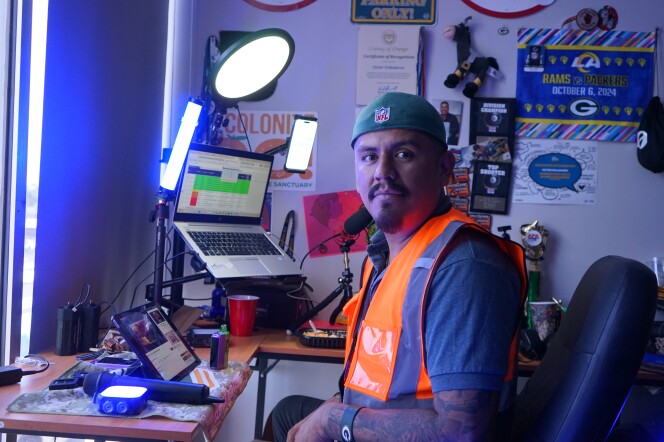 A man with a medium skin tone, a mustache and goatee sits in an office chair. He wears a green baseball hat backwards, and an orange vest over a gray t-shirt. Behind him, there's a laptop, phone and lights set up on a desk.