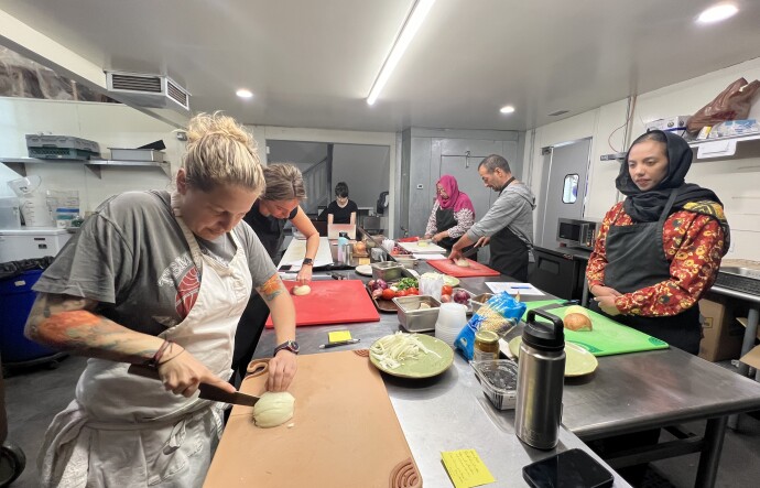 Four people of various shades stand around in a kitchen cutting onions. 