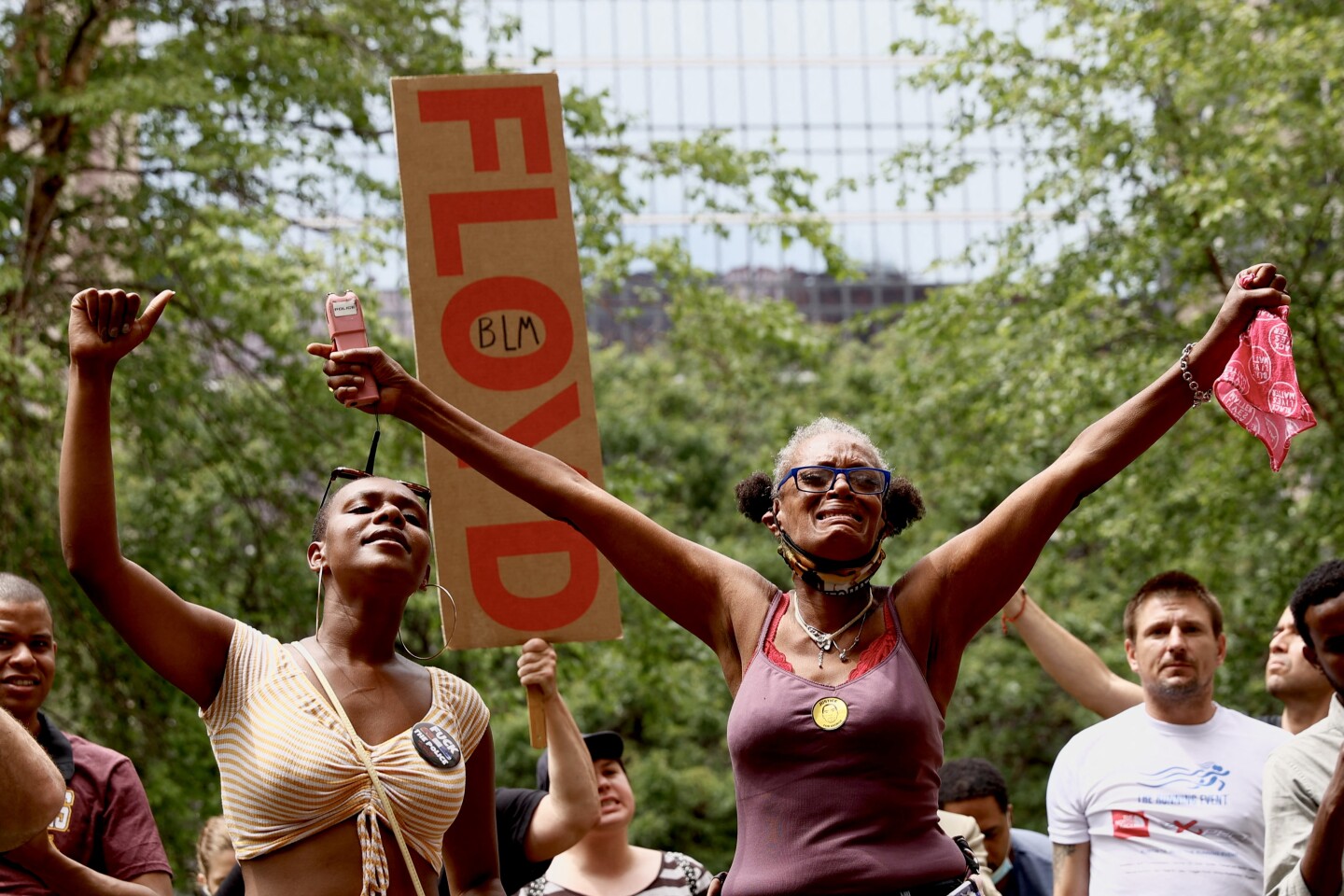 A woman in tears raises her arms, backed by a small crowd, with one person raising a sign with the words "Floyd" and "BLM."