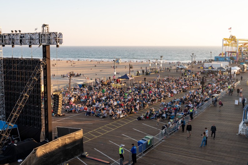 A crowd of people are gathered for a screening on the Santa Monica Beach Pier. Behind the audience, the pier rides and ocean are visible.