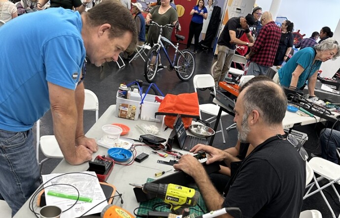A room full of men and women of all ages. A white foldable table is set up closer to the camera, with two people seated behind it, including a man in blue jeans and a black t-shirt. Another man wearing a blue t-shirt and jeans is leaning over the table, which is covered in tools and electrical equipment.