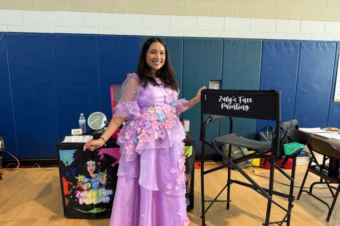 A brown skinned woman in a purple tulle dress next to a chair that says "Zuly's face painting" in a gym. Her art supplies are in the background.