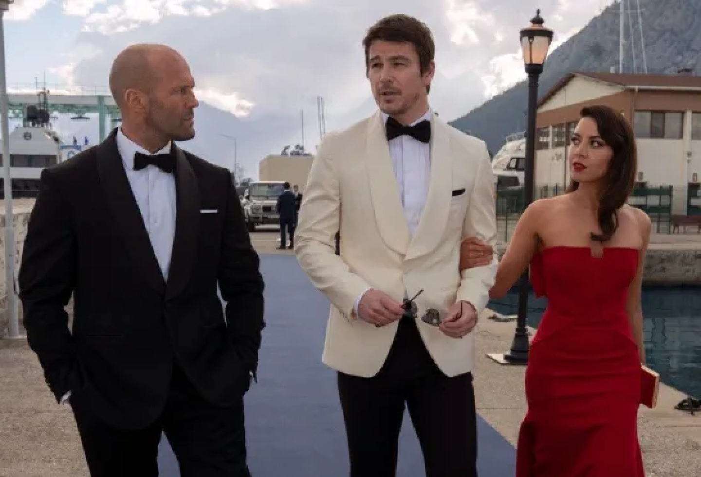 Jason Statham, Josh Hartnett, and Aubrey Plaza walk side by side along a dock of a harbor. Large yachts can be seen behind them. Statham is to the left, wearing a black suit with a white button-up shirt and black bow-tie. He looks at Hartnett who is next to him on the right. Hartnett wears black pants with a white suit jacket and black bow-tie. He is holding sunglasses at his waist in front of him with both hands. He looks towards Statham seemingly mid sentence. Plaza is to Hartnett's right, holding onto his arm closest to her. She wears a red strapless dress that is fitted. It matches the red lipstick she is wearing and the small clutch she holds on in her left hand. She looks over to Hartnett as well.