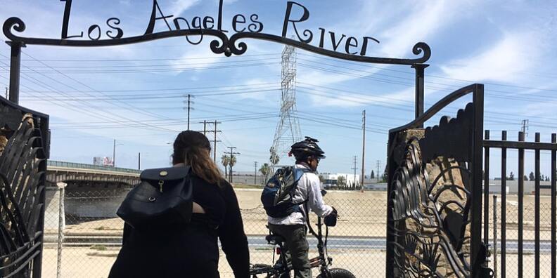 Two people on bicycles ride through a metal gate with a sign that reads "Los Angeles River." The river can be seen in the background, below them.