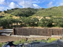 A wall with scaffolding stands in front of a hillside lined with trees and grass.  