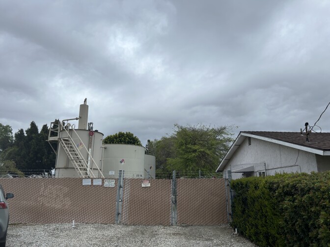 An off white oil holding tank next to a single-family home under a cloudy sky. A tan fence surrounds the oil tank.  