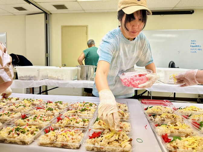 An Asian American woman wearing a baseball cap holds a large tupperware full of ginger. She leans over to place some on top of one of the plates of rice lined up on the table.