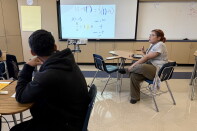 A teenage boy with medium skin tone and a black hoodie looks at a projection of math calculations. A women with medium light skin tone and glasses sits at a desk and gestures as though mid-speech. 