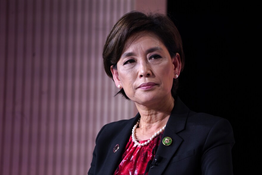 A woman with short black hair cut just above her ears is looking to her right, with her head slightly angled towards the camera. She's wearing a black blazer, red shirt, pearl necklace and a green pin on her right side.