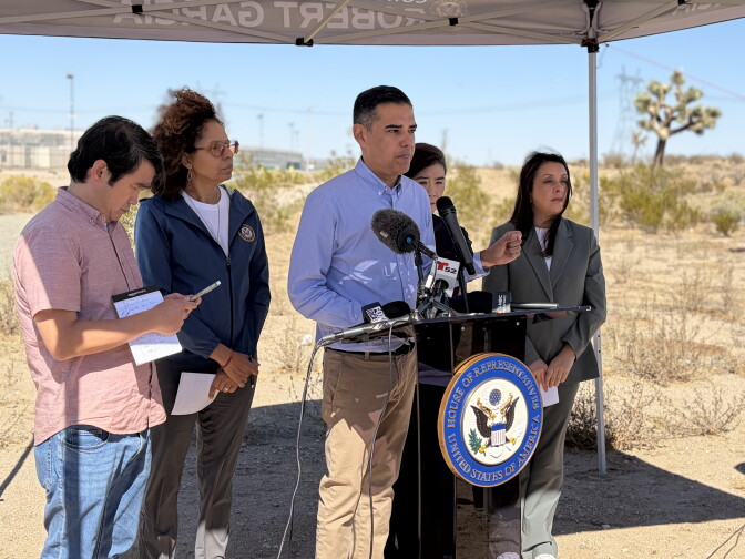 A man speaks at a House of Representatives podium outdoors in the desert, with four others standing nearby under a tent.