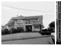 A black and white wide shot of a home from the street on the left. On the right is a view of a woman from the ground up smiling while standing in front of a jail cell's doors.