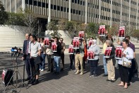 Two people stand behind a portable mic stand, one is clad in a suit and tie, the other has lifted their pant leg to reveal an ankle monitor. Behind them, about a dozen people hold up red, black, and white signs that read: "Drop All Charges Against John"