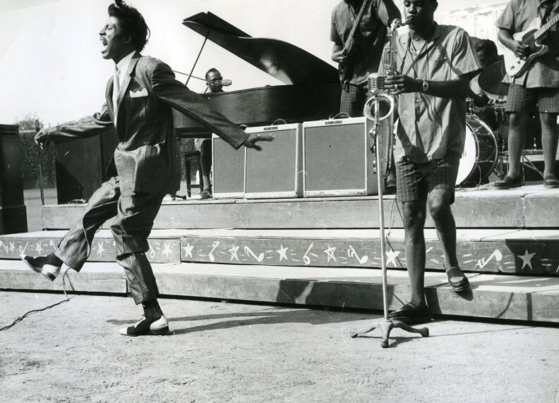 Little Richard dances on stage in a suit, energetic, with an all-Black band behind him playing guitar, bass, saxophone, drums, and piano.