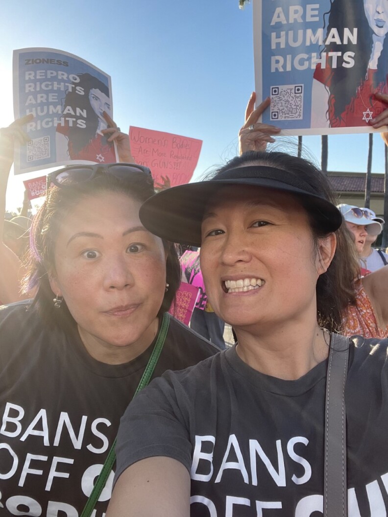 Two Asian American women wear black shirts at a rally for abortion rights. People in the background hold signs that read "Repro Rights are Human Rights."