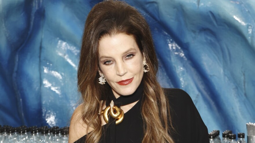 A light-skinned woman with long brown hair, wearing red lipstick and a black dress, stands in front of a blue background.