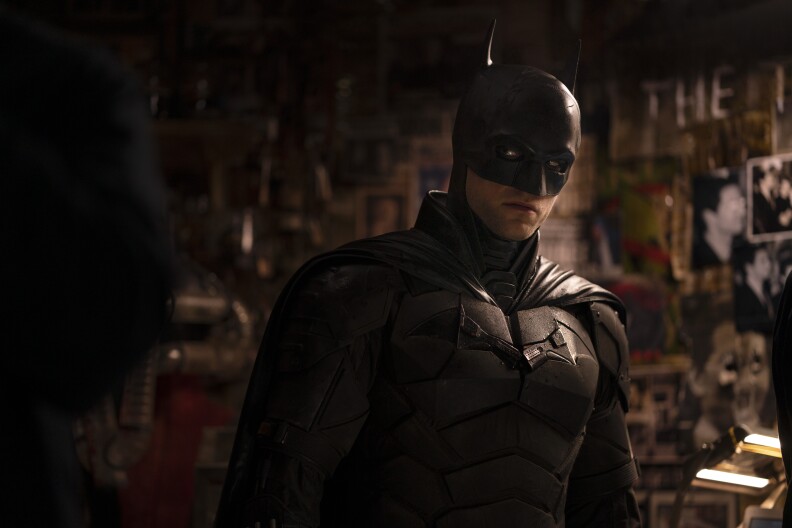 A man in a thick coat with a scarf talks and points, while another man in full Batman costume stands by.