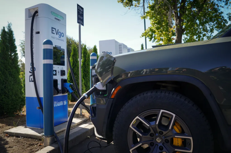 A black electric SUV is charging at a white and blue station with text on it that reads "EVgo."